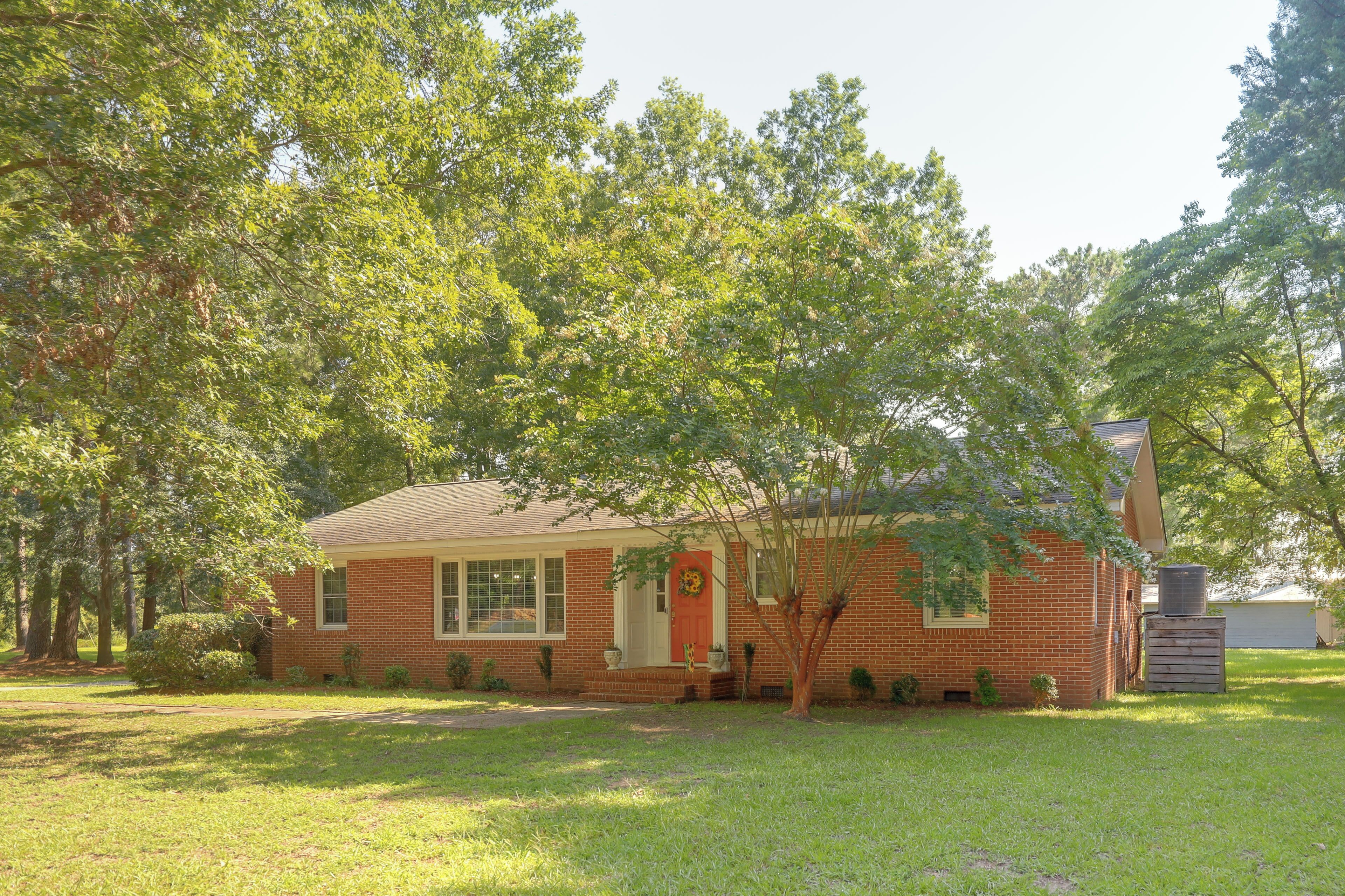 A single-story brick house with a front porch, a small garden, and a tall tree in front.