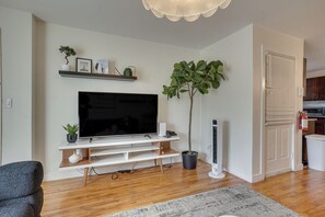 A living room with a flat-screen TV on a white entertainment unit, a potted plant, and a fire extinguisher mounted on the wall. - Easy NYC Commute: Furnished Jersey City Home (Jersey City)