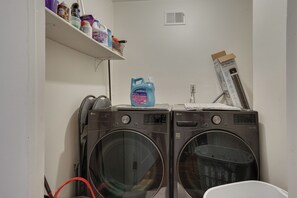 A laundry area with two front-loading washers, a shelf with cleaning supplies, and a vent on the wall. - Easy NYC Commute: Furnished Jersey City Home (Jersey City)