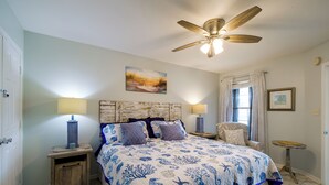 Une chambre à coucher comprenant un lit, deux tables de chevet, un ventilateur de plafond, une fenêtre avec des rideaux et un tableau accroch é au mur.