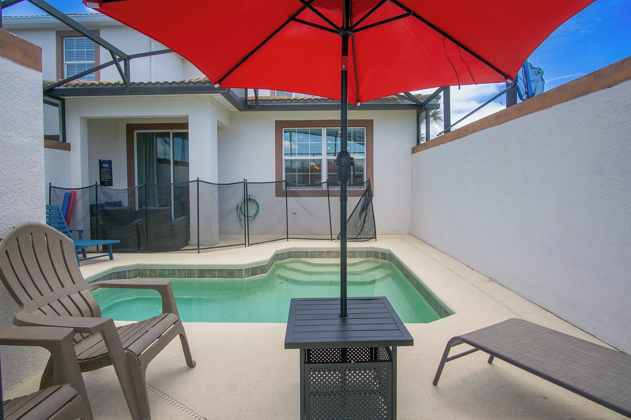 A swimming pool with a red umbrella, a chair, and a table.