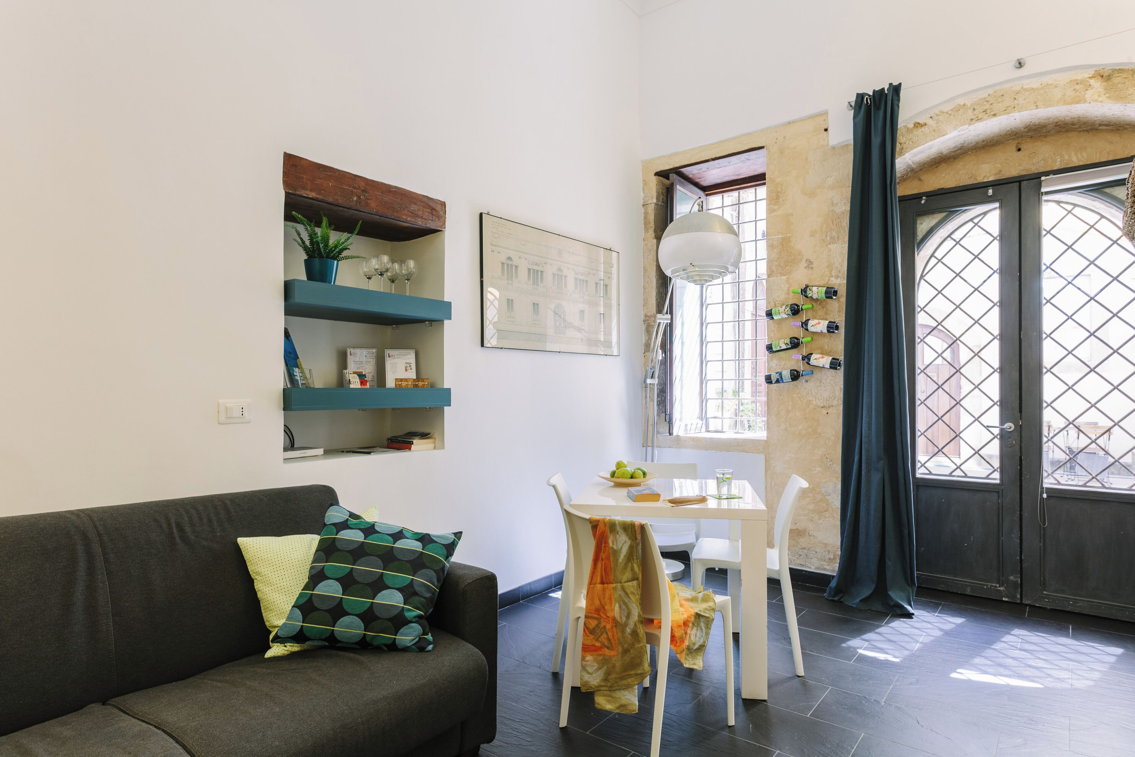 A modern living room with a sofa, a dining table with chairs, a window with curtains, and a wall-mounted shelf with decorative items.
