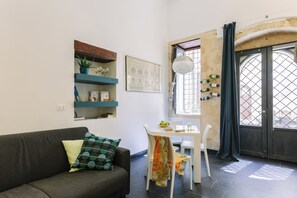 Ein modernes Wohnzimmer mit einer Couch, einem Esstisch mit Stühlen, einem Fenster mit Vorhängen und einem Wandregal mit Dekoration.