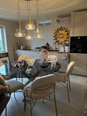 A modern kitchen with a dining area, featuring a glass-top table, white chairs, and pendant lighting.