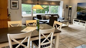 A dining area with a table and chairs, a sofa, a television, and a window with a view of greenery. - Süderstraße 29a, Apt. 01 - Spacious 3-room vacation apartment with a terrace and (Sylt)