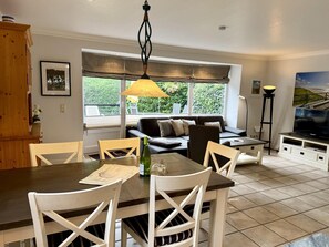 A dining area with a table and chairs, a sofa, a television, and a window with a view of greenery. - Süderstraße 29a, Apt. 01 - Spacious 3-room vacation apartment with a terrace and (Sylt)