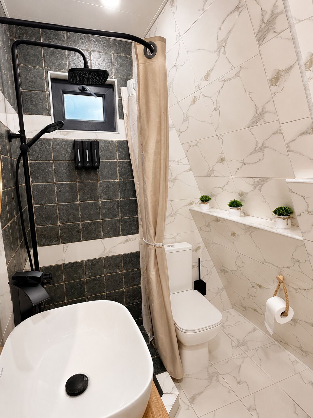 Une salle de bain avec un lavabo blanc, une douche avec un rideau beige, des toilettes et une étagère avec des plantes.