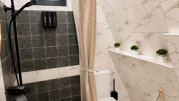 Une salle de bain avec un lavabo blanc, une douche avec un rideau beige, des toilettes et une étagère avec des plantes.