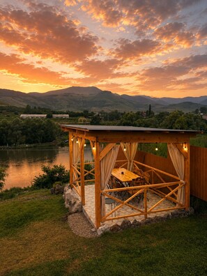 A wooden gazebo with a table and chairs, surrounded by a stone wall, overlooking a lake and hills.