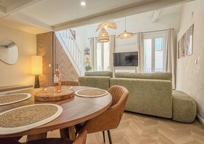 Un salon moderne avec un coin repas, comprenant une table en bois, des sets de table en fibres naturelles, un canapé et un téléviseur.