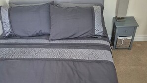 Una cama con ropa de cama gris, una mesita de noche con una lámpara y una pequeña cesta de mimbre.