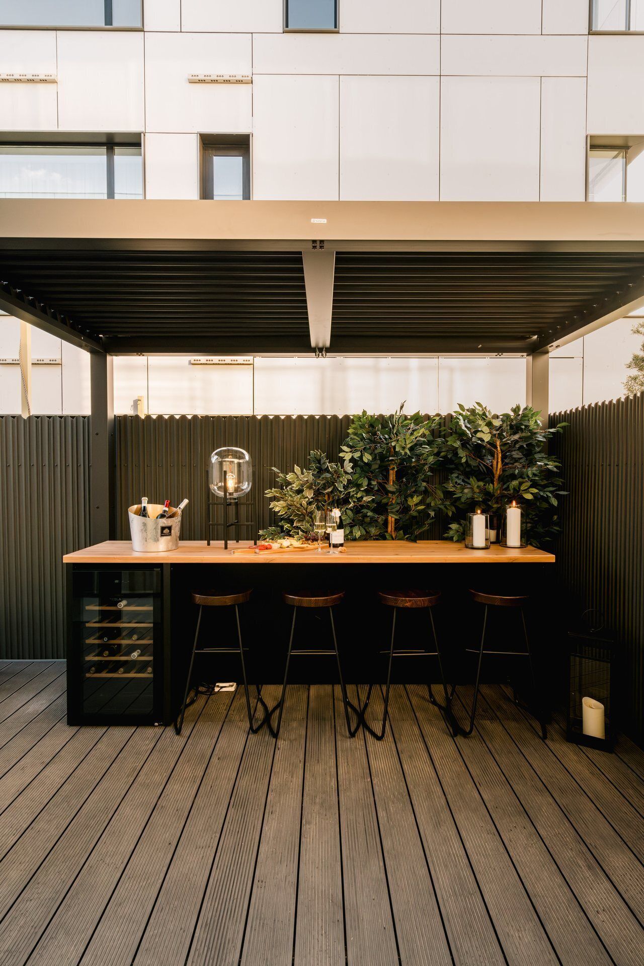 Un área de bar al aire libre con techo, una mesa de madera, taburetes de bar, un enfriador de vino y una planta grande en maceta.