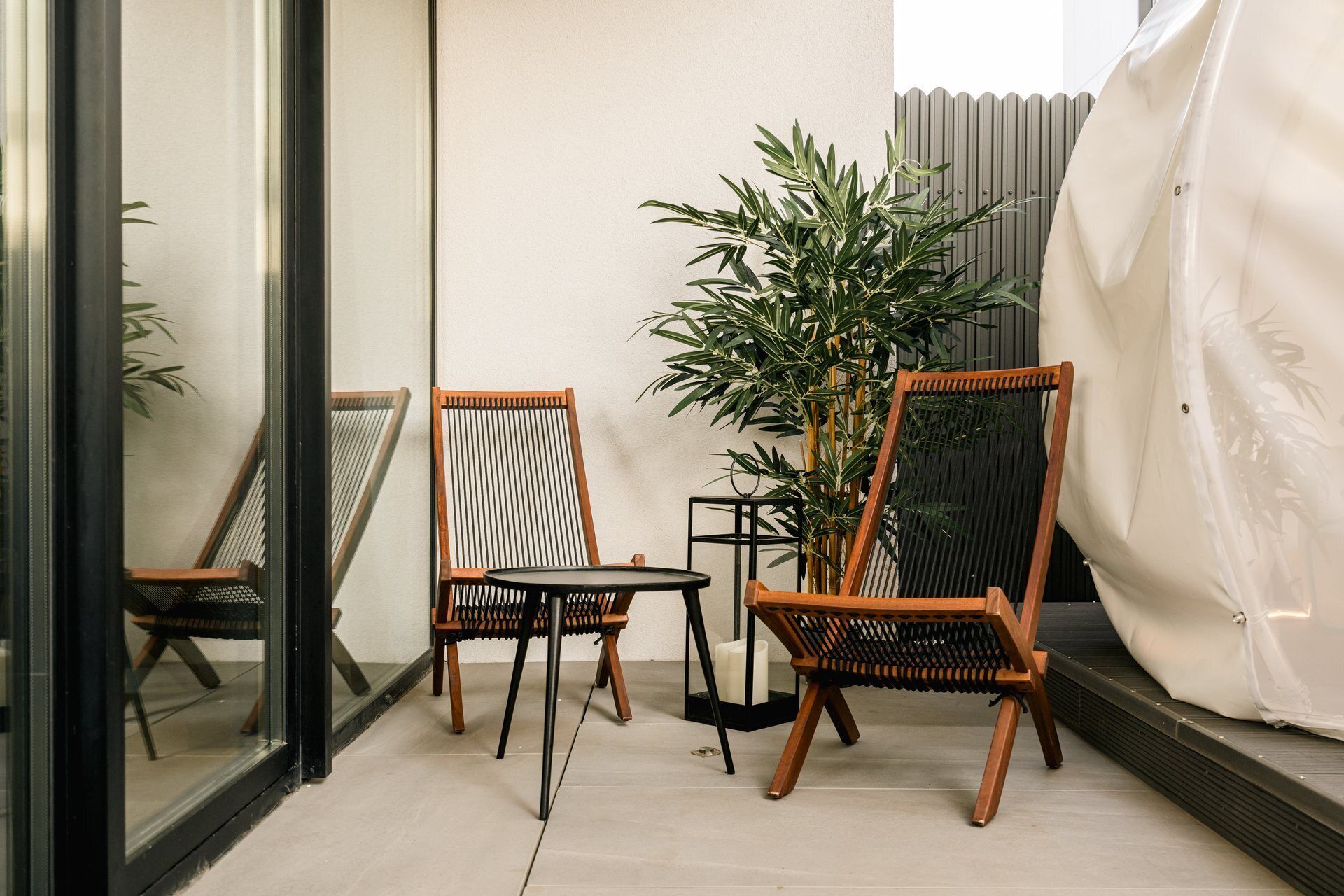 Un petit espace extérieur aménagé avec deux chaises en bois et une table, entouré de plantes en pot et d’un rideau blanc.