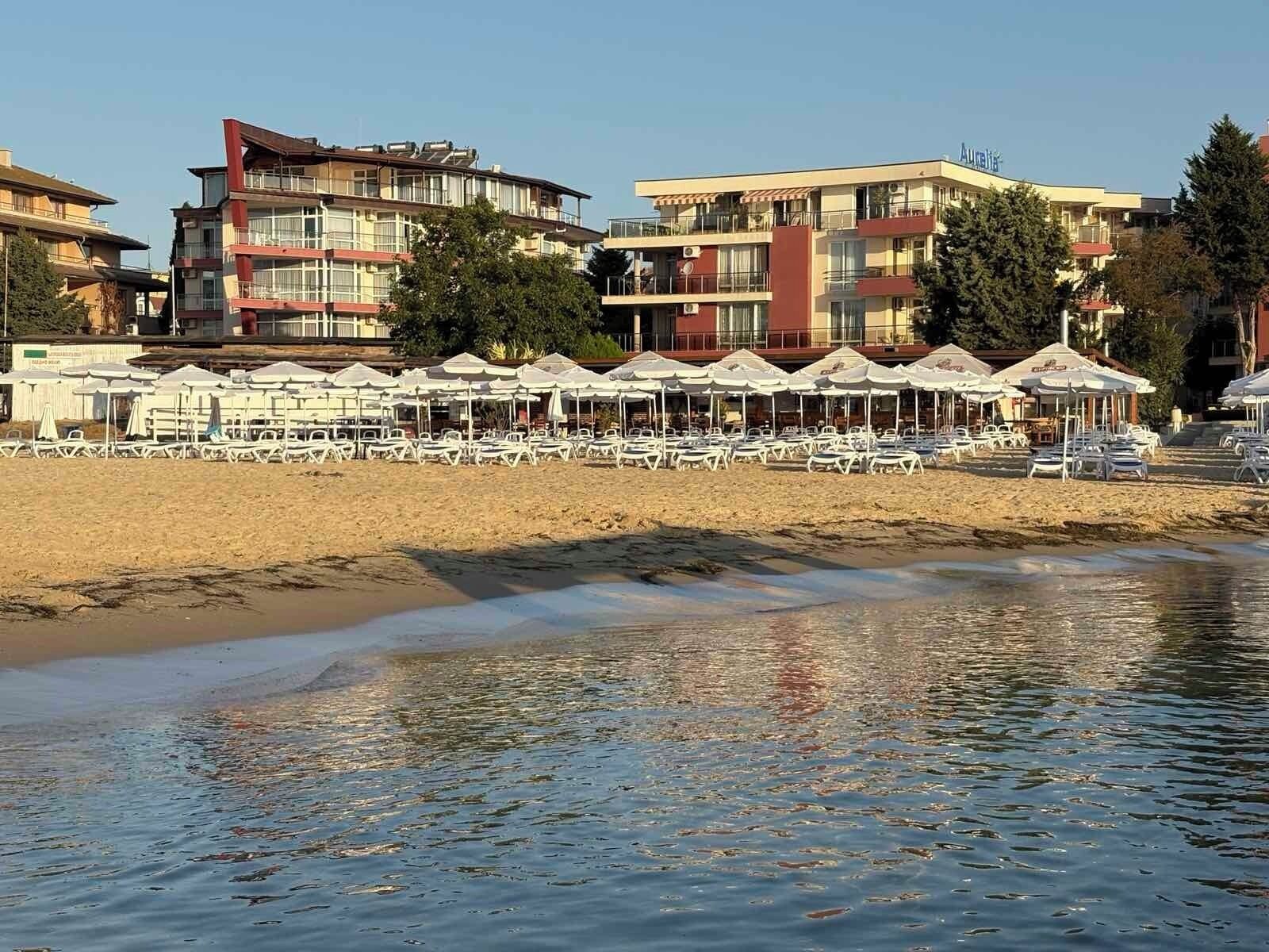 A beachside hotel with multiple-story buildings, outdoor seating with umbrellas, and a sandy beach.