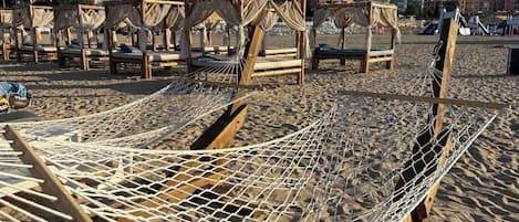 A beach with hammocks and wooden chairs, a clear sky, and buildings in the background.
