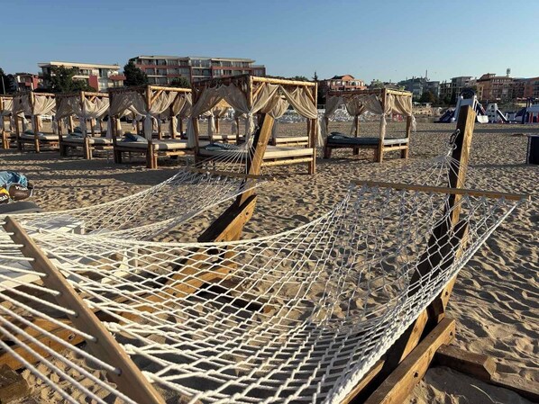 A beach with hammocks and wooden chairs, a clear sky, and buildings in the background.