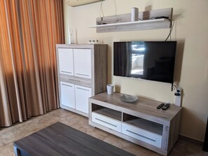 A room with a flat-screen TV mounted on the wall, a wooden cabinet with drawers, a small table with a cup and remote controls, and a window with curtains. - Komfortables Apartment mit 2 Schlafzimmern direkt am Strand in Nessebar (Nesebar)
