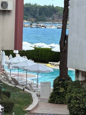 A pool area with lounge chairs and umbrellas, a sign for 'Aurelia', and a view of the sea with boats. - Komfortables Apartment mit 2 Schlafzimmern direkt am Strand in Nessebar (Nesebar)
