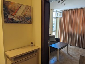 Ein Zimmer mit einem Segelbootgemälde, einem kleinen Tisch mit einer Flasche und Blick durch ein mit Vorhängen versehenes Fenster nach draußen.