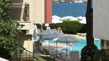 A pool area with lounge chairs and umbrellas, a view of the sea, and a building with balconies.