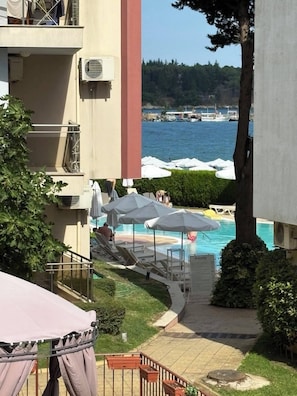 A pool area with lounge chairs and umbrellas, a view of the sea, and a building with balconies.