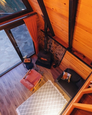 Interior de una cabaña de madera con chimenea de piedra, un sofá, una silla y una pequeña mesa con un jarrón de flores.