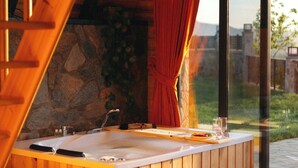 Una bañera de madera con un muro de piedra de fondo, una ventana con cortinas rojas y vistas a la vegetación exterior.