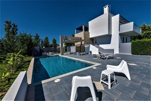 Un área de piscina moderna con una piscina rectangular, sillones de descanso y un edificio de dos plantas con fachada de piedra.