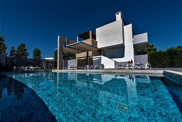 Un área moderna junto a la piscina con una piscina azul cristalina, sillones blancos y un edificio contemporáneo con detalles de piedra.