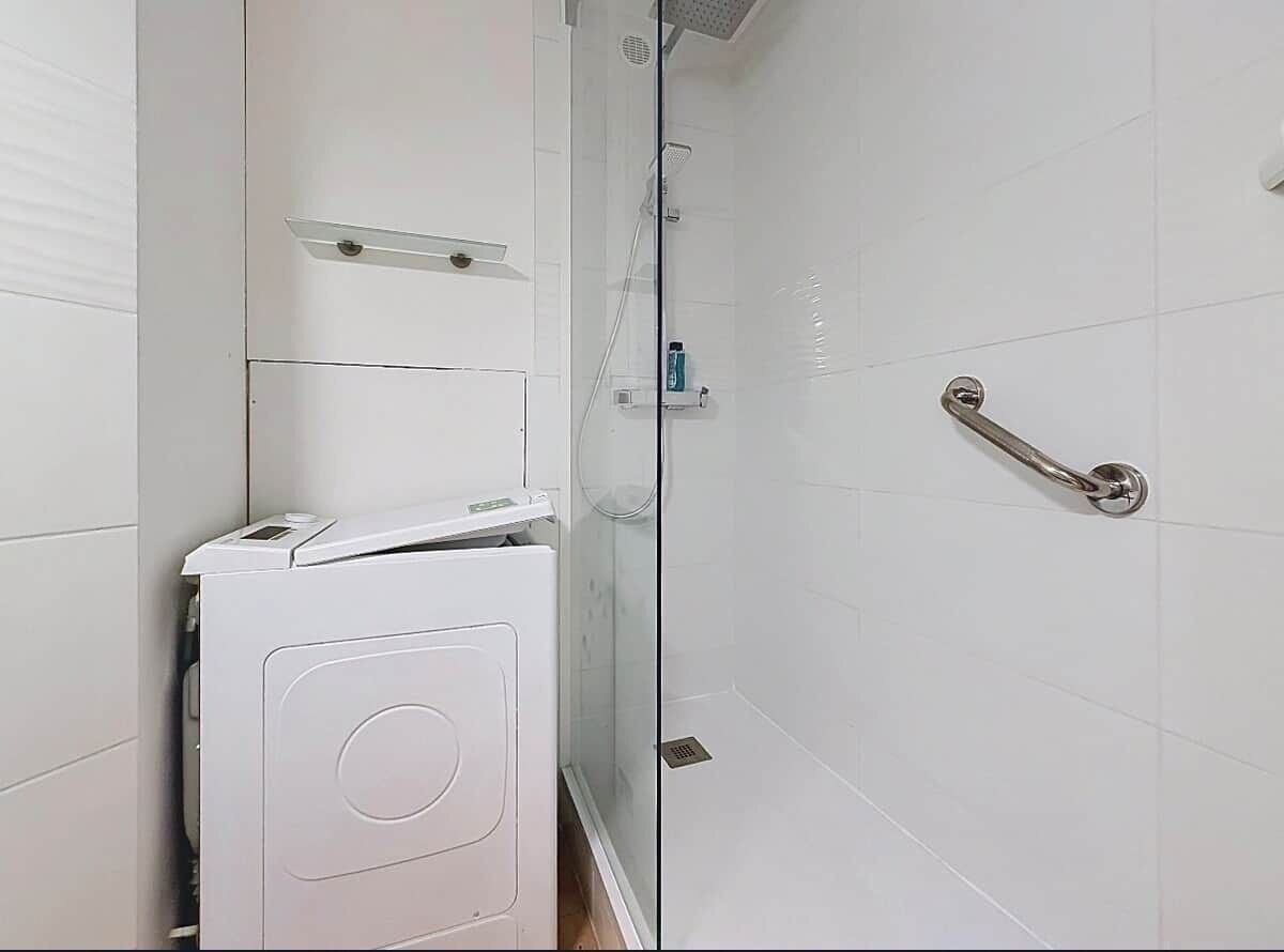 Un baño con pared de azulejos blancos, cabina de ducha de cristal, lavadora blanca y barandilla metálica.