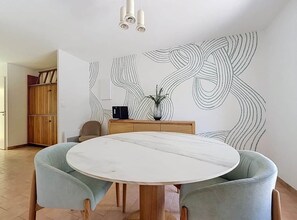 Ein Essbereich mit einem runden Tisch, zwei Polsterstühlen, einer Holzkommode und einer Wand mit einem großen abstrakten Muster.