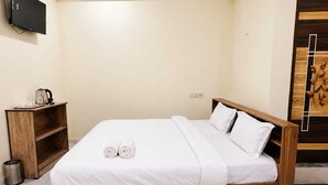 A neatly made bed with white linens and pillows, a wooden bedside table with a kettle, and a television mounted on the wall.