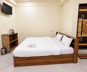 A neatly made bed with white linens and pillows, a wooden bedside table with a kettle, and a television mounted on the wall.