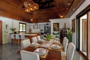 A modern kitchen with a wooden ceiling, a dining area with a large table set for a meal, and a view of the outdoors through a sliding door. - Surin Lake Villa (Thalang District)