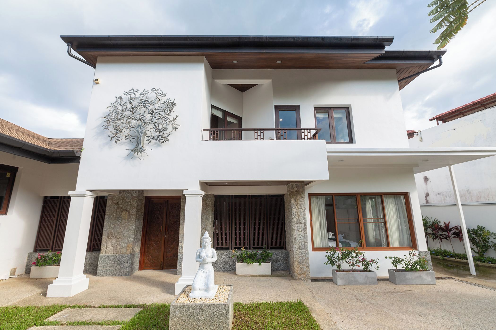 Une maison à deux étages avec un balcon, une sculpture d’arbre décorative et une statue dans le jardin avant.