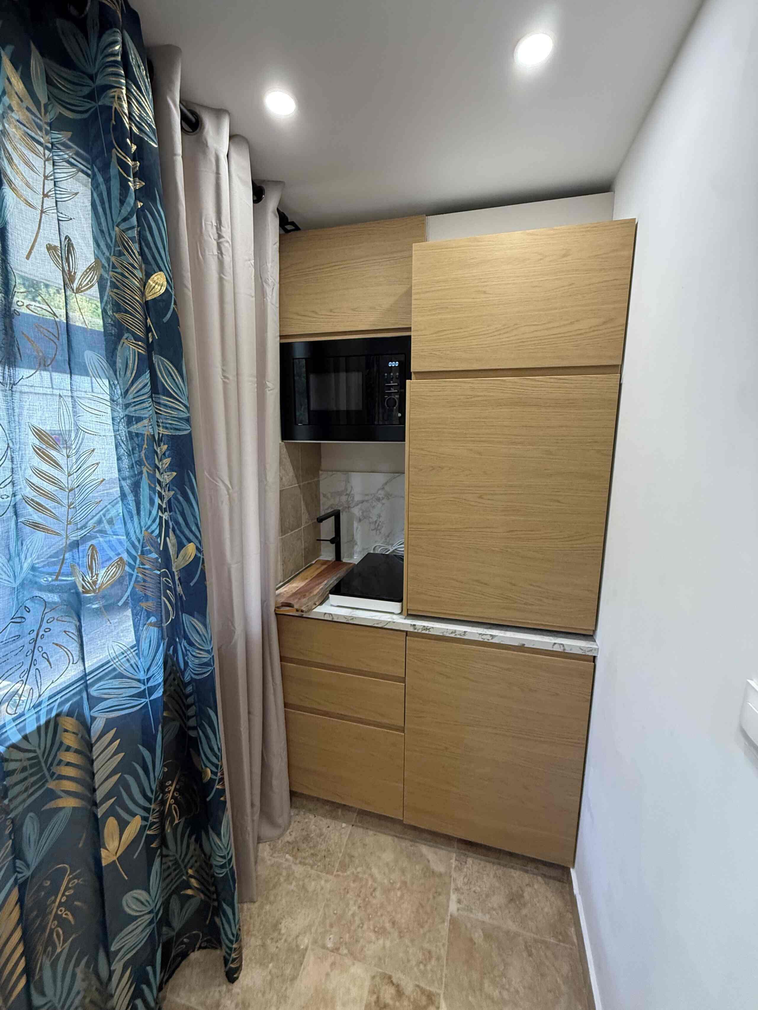 A compact room with a kitchenette, a bed, and a curtain with a leaf pattern.