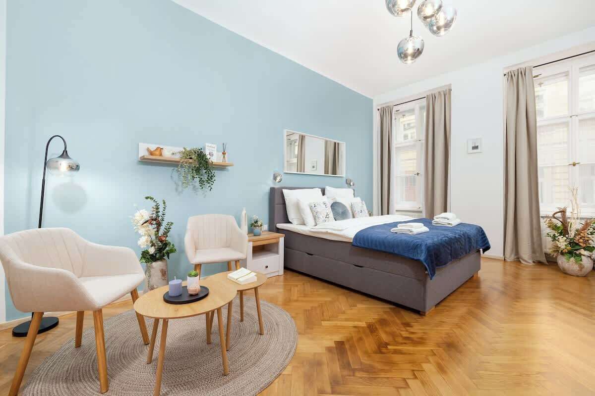 A modern bedroom with a bed, two chairs, a small table, a bedside table, a mirror, and a window with curtains.