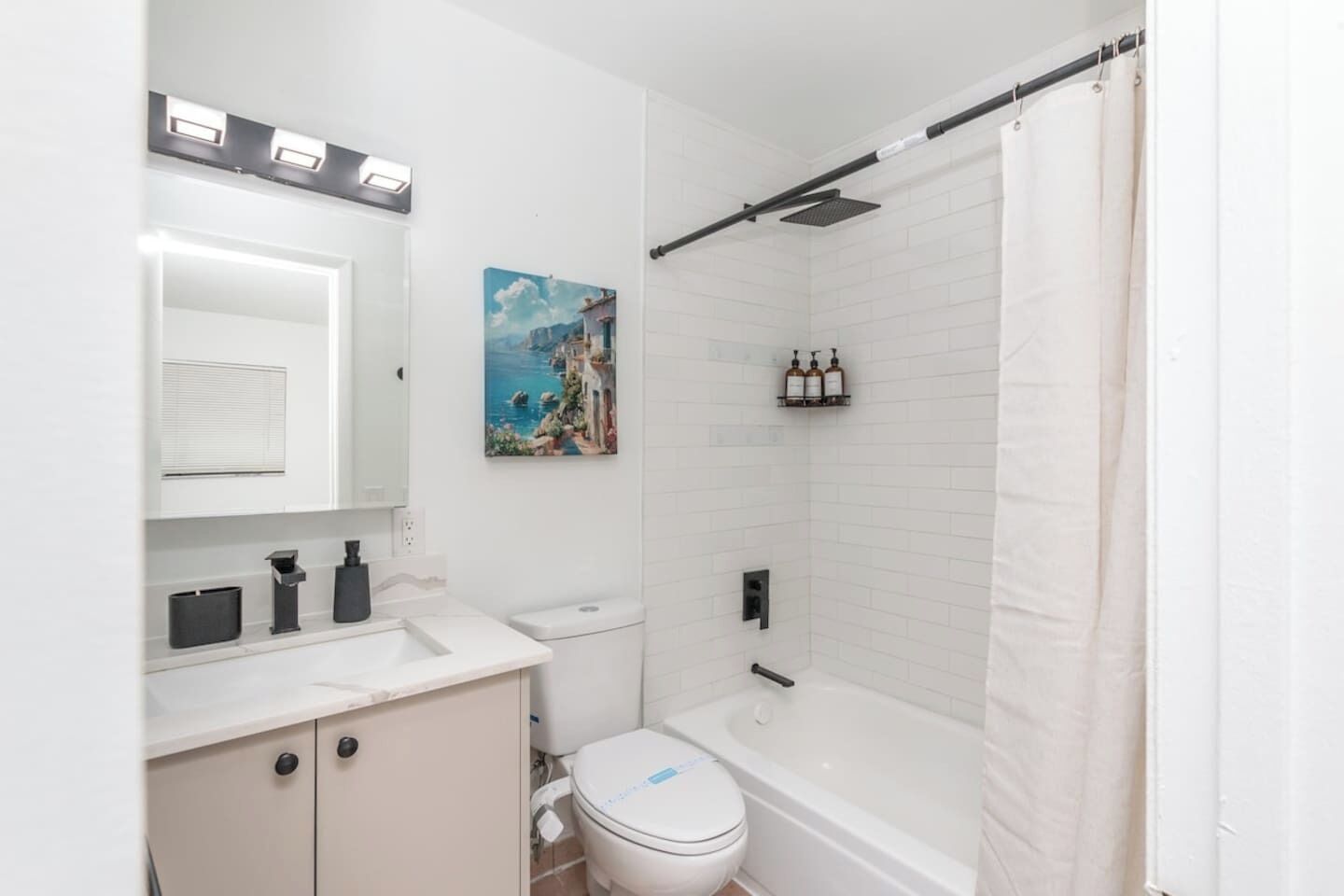 A modern bathroom with a white sink, toilet, and bathtub. There is a shower curtain, a painting on the wall, and a shelf with toiletries.