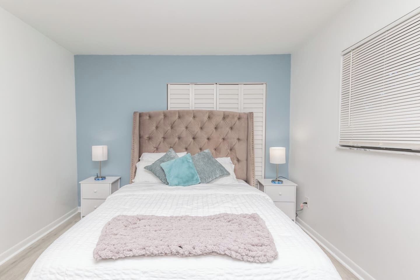A bedroom with a tufted headboard, white bedding, two bedside lamps, and a window with blinds.