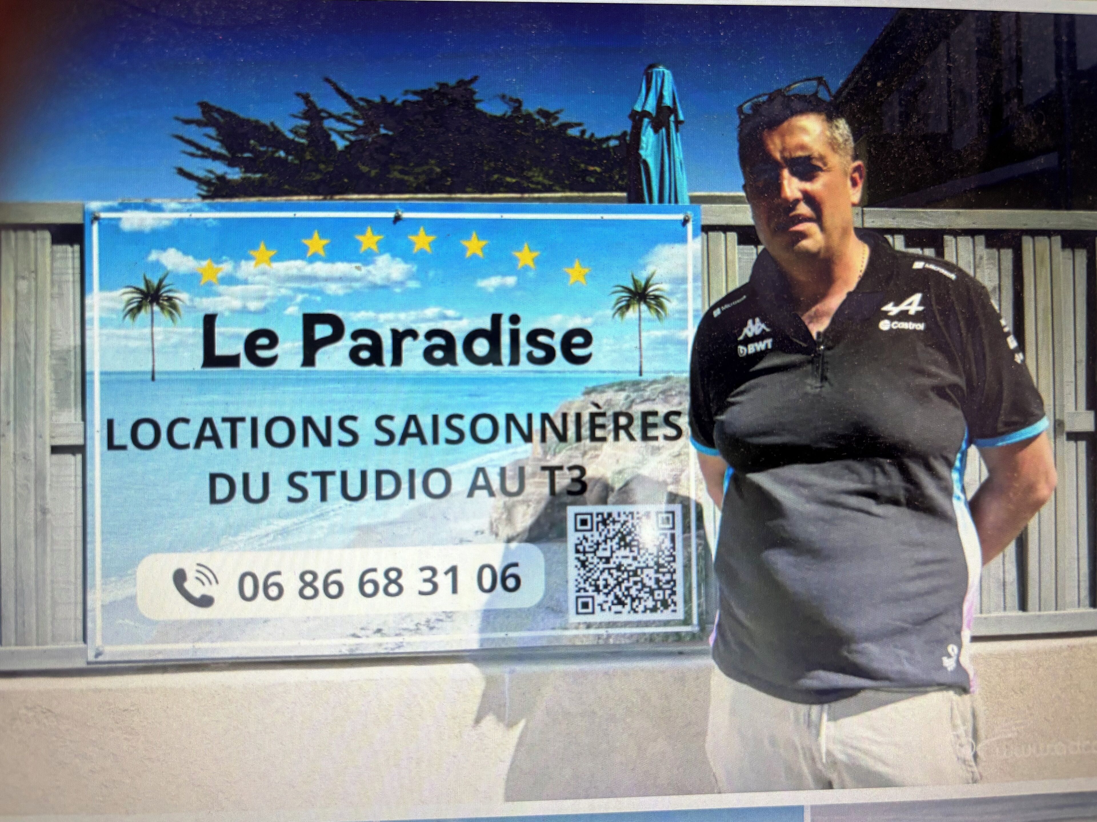 A man standing in front of a sign advertising 'Le Paradise' with a beach scene, a phone number, and a QR code.