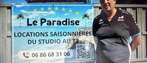 En man står framför en skylt som annonserar för ”Le Paradise”, och på skylten syns en strandvy, ett telefonnummer och en QR-kod.