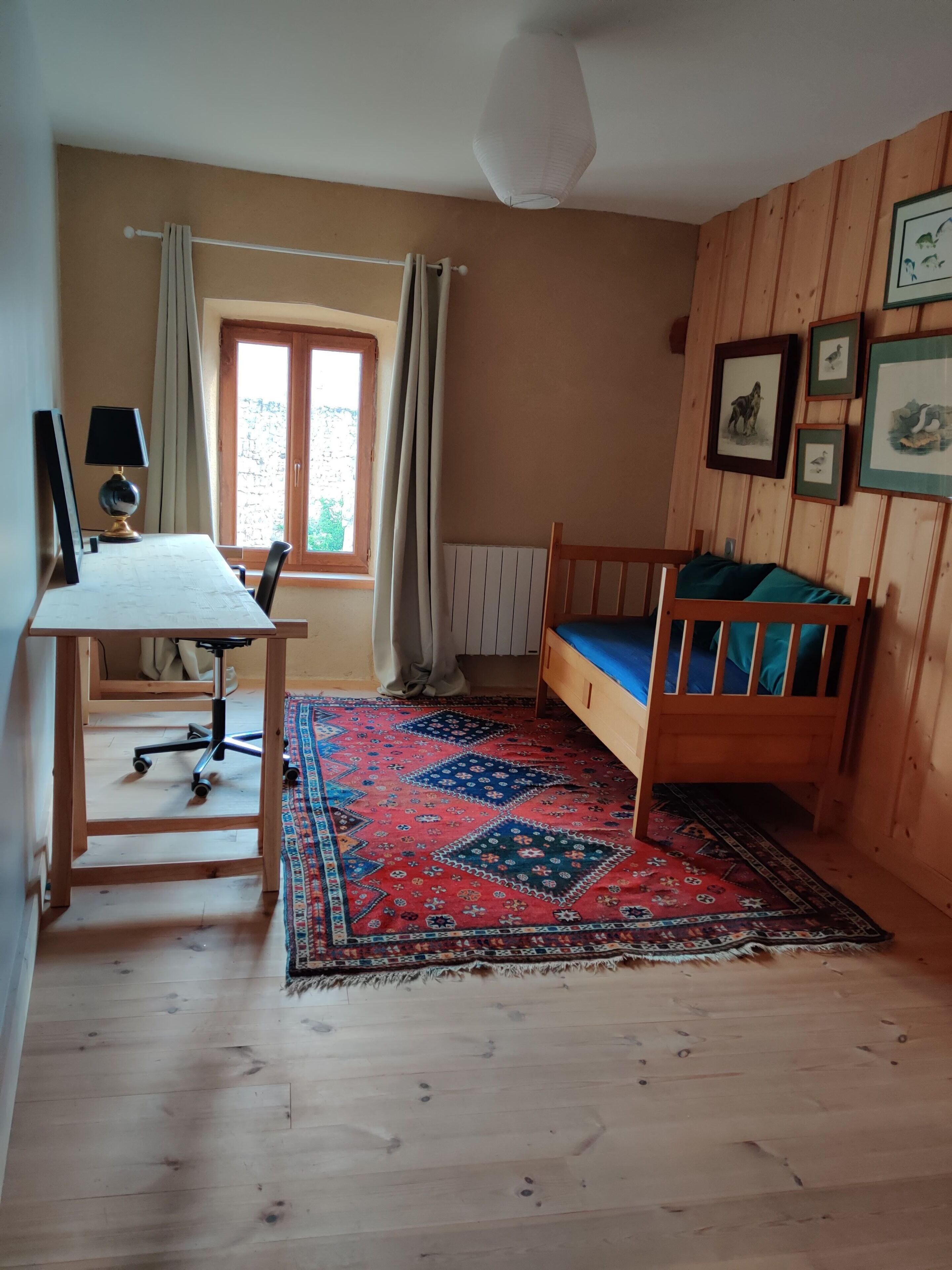 Une pièce avec un lit en bois, un bureau avec une chaise, une fenêtre avec des rideaux et un tapis à motifs.