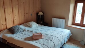 A bedroom with a wooden floor, a bed with a white and patterned bedspread, a bedside lamp, a window with a view, and a radiator.
