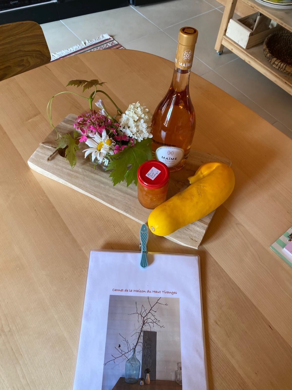 A wooden table with a bottle of wine, a jar, a yellow squash, and a piece of paper with text.