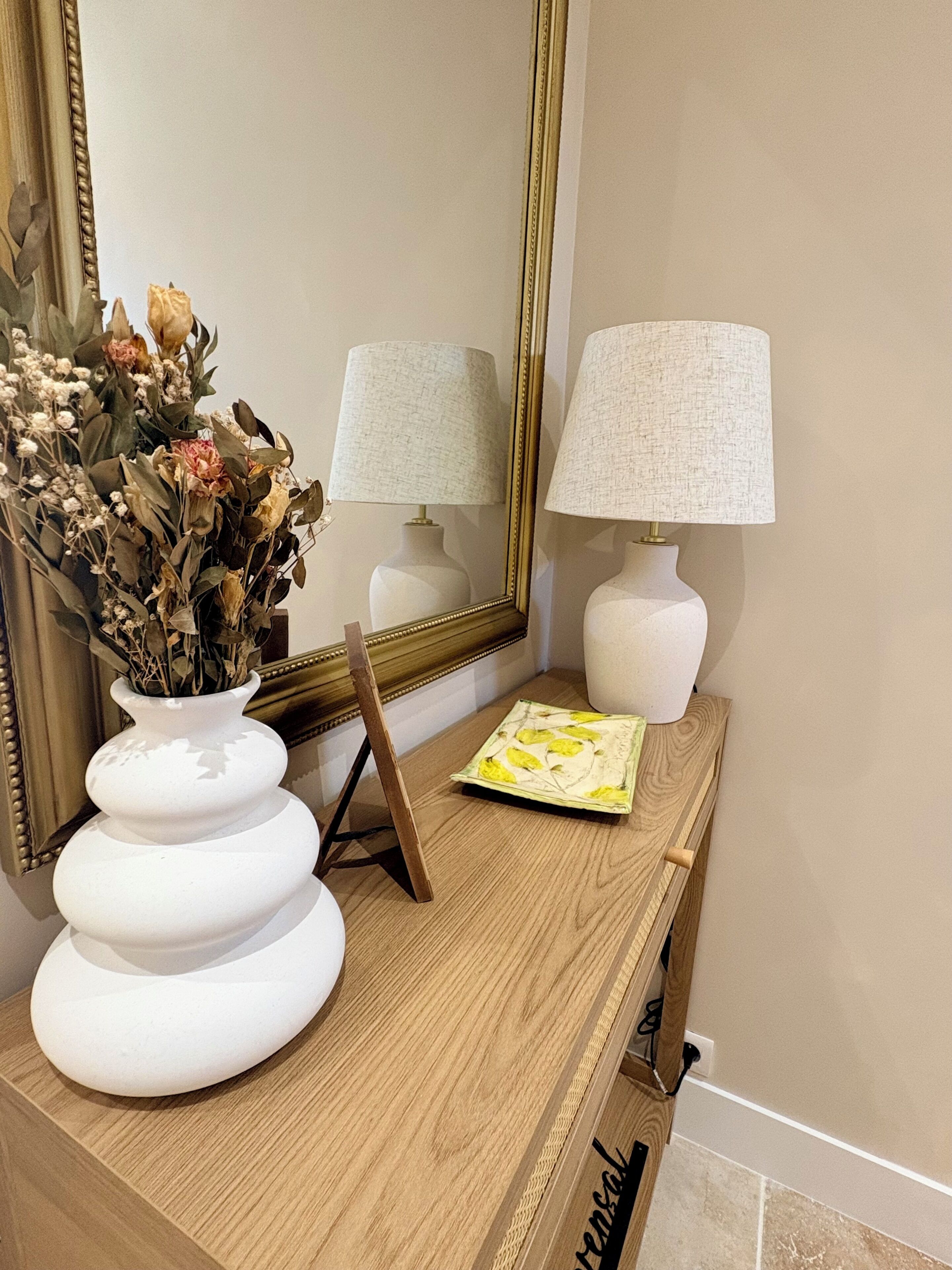 Una consola de madera con un jarrón blanco de flores secas, dos lámparas blancas y un espejo enmarcado.