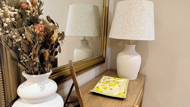 Une console en bois surmontée d’un vase blanc contenant des fleurs séchées, de deux lampes blanches et d’un miroir encadré.