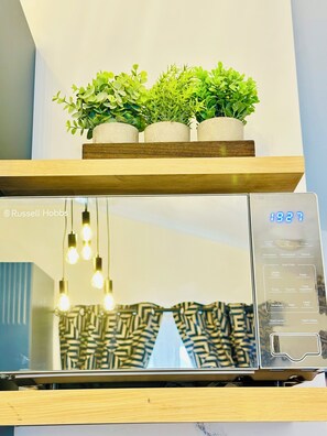A microwave with a digital display, a wooden shelf with potted plants, and hanging light bulbs. - Modern Self-Contained Studio Annexe with Kitchenette and Private Entrance (Oxford)