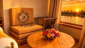 Una stanza con vista su un tramonto sull'acqua, un tavolo di legno con un centrotavola floreale e un'area salotto con cuscini decorati.