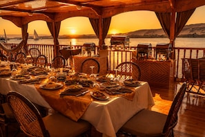 A dining area with a view of the sunset over water, equipped with tables, chairs, and a barbecue setup.