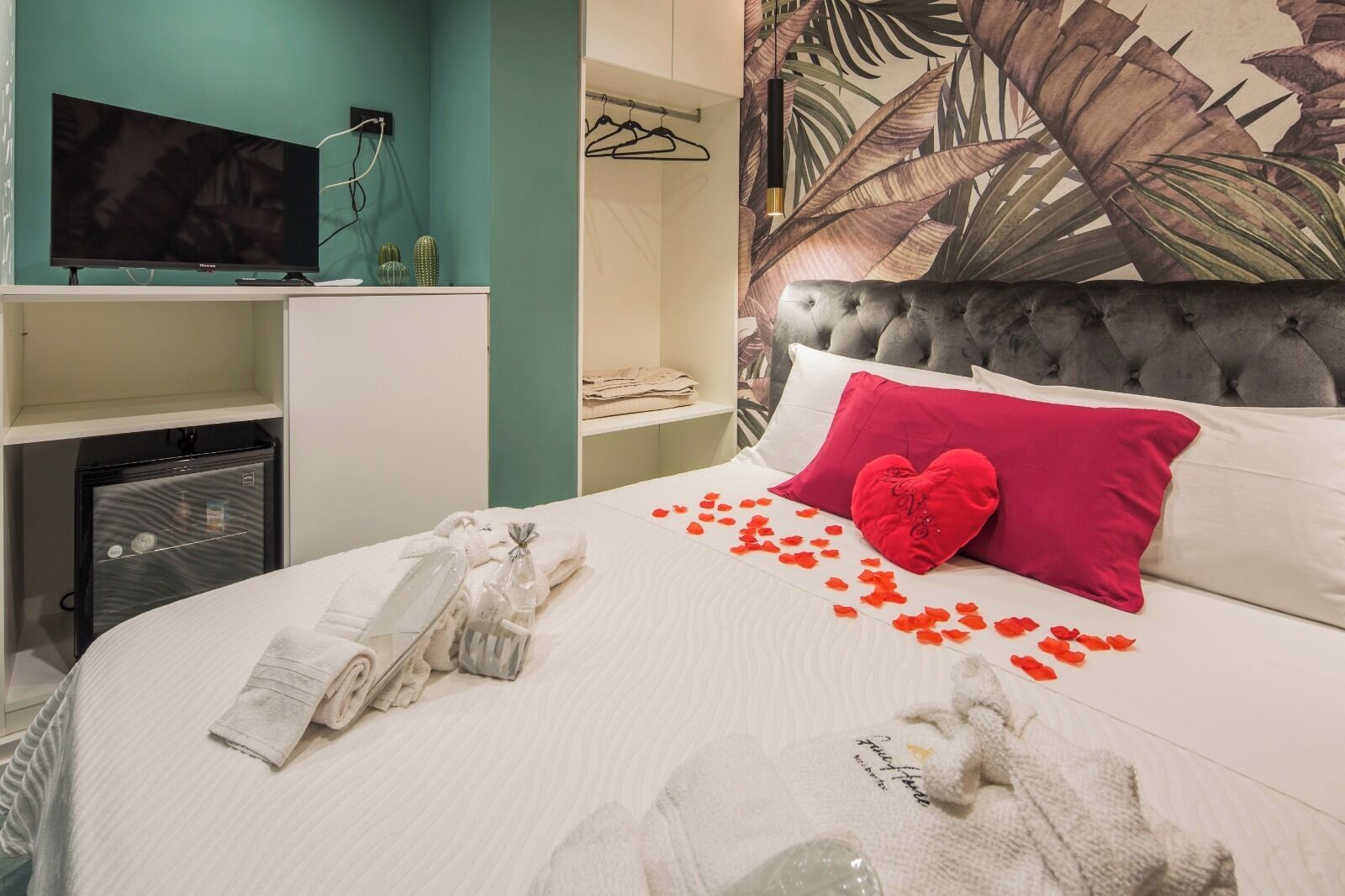 Habitación de hotel con cama, televisión, mini bar y un mural de hojas tropicales en la pared.